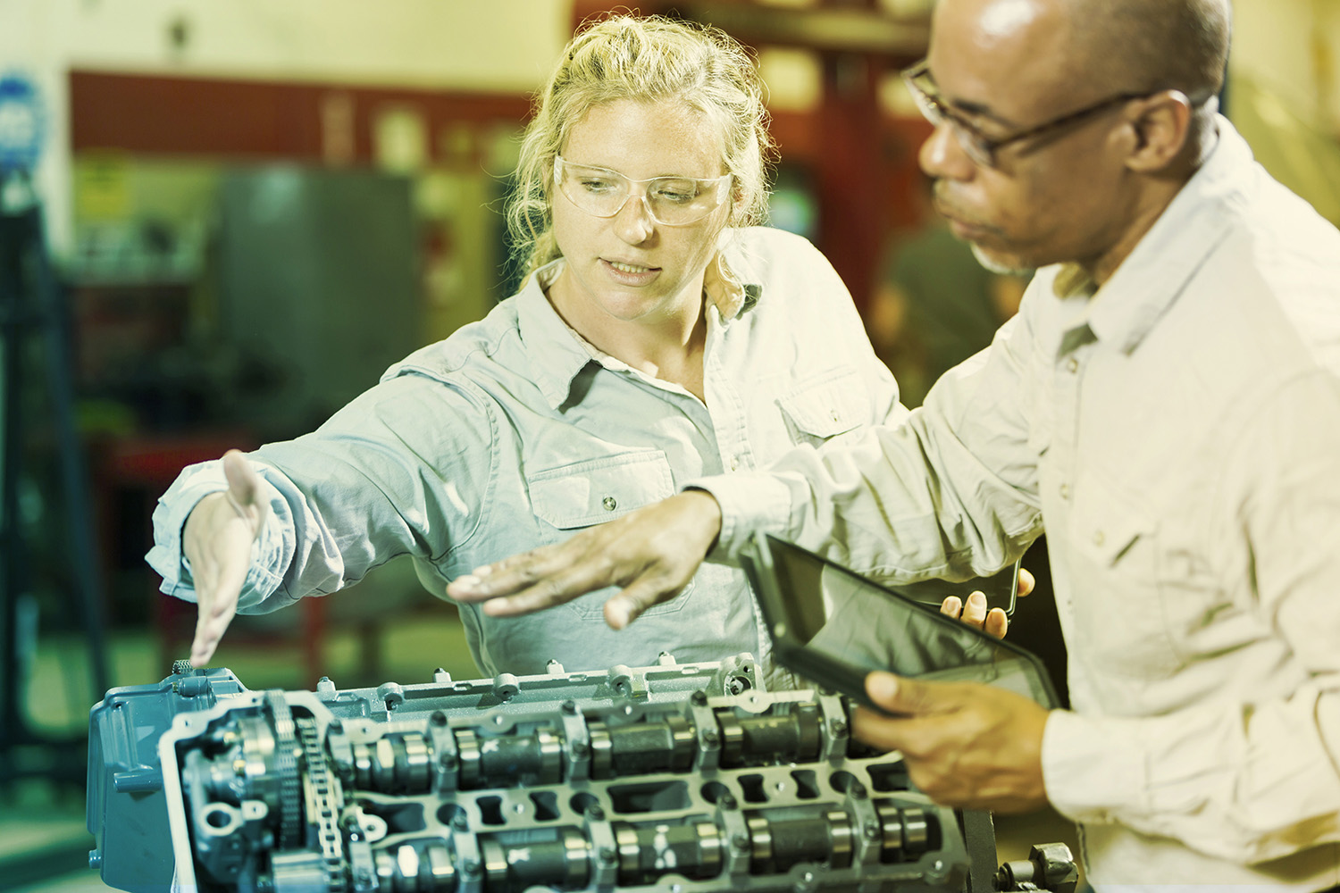 Two multi-ethnic mechanics working on a gasoline engine in a repair shop.  The African American man, in his 50s, is explaining to the trainee, a woman in her 20s, how to repair the engine.  He is holding a digital tablet.  The focus is on the woman